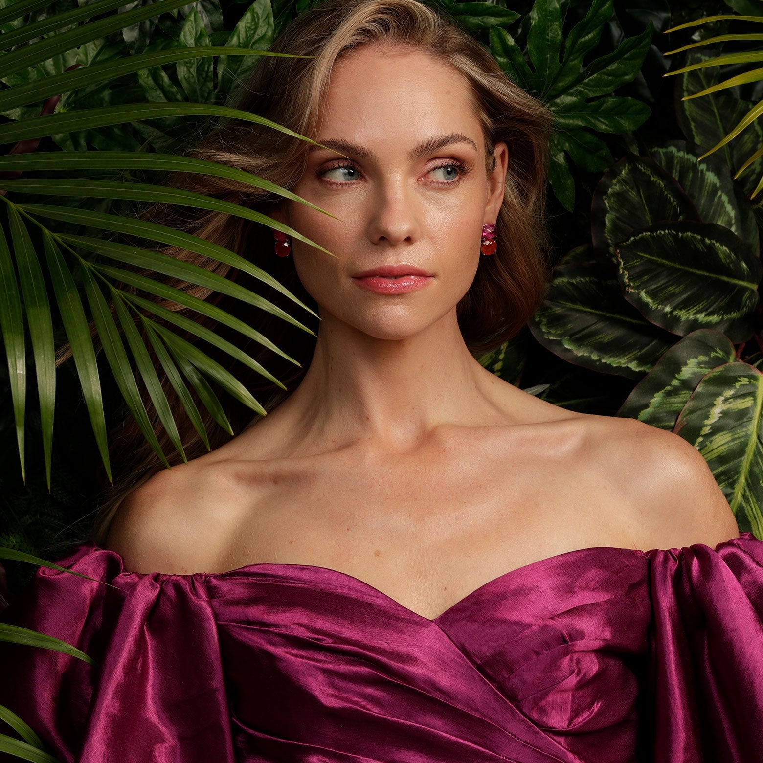 A woman in a deep magenta off the shoulder top stands in front of a green wall. She is wearing the Kelly Woodcroft bottle brush earrings earrings with pink and red oval shaped stones set in silver