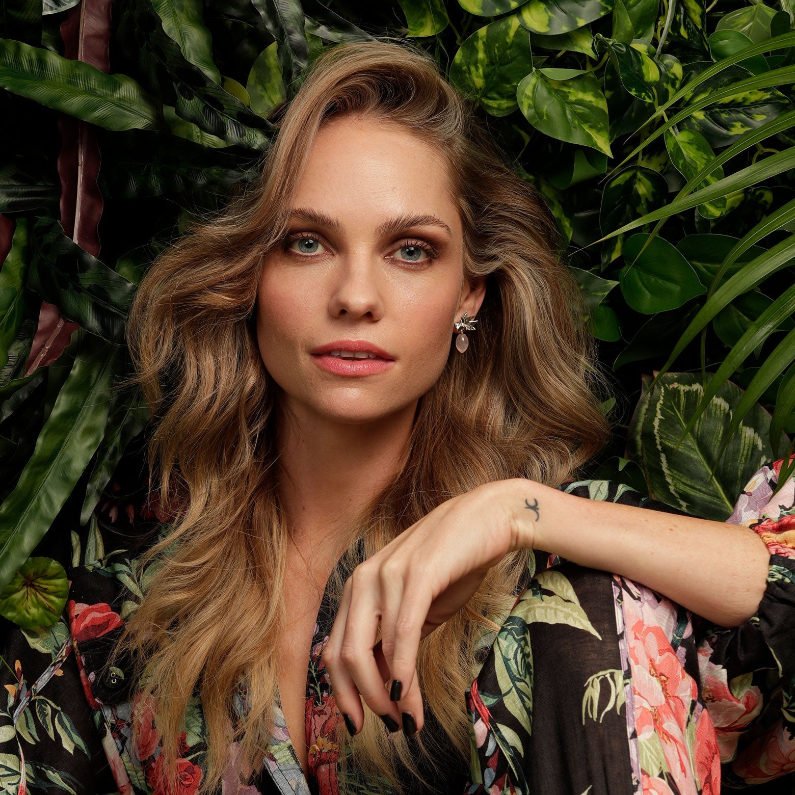 A woman in a black and floral print jumpsuit sits in front of a green wall. She is wearing the Kelly Woodcroft orchid earrings that feature a silver flower motif and quartz coloured dangle.